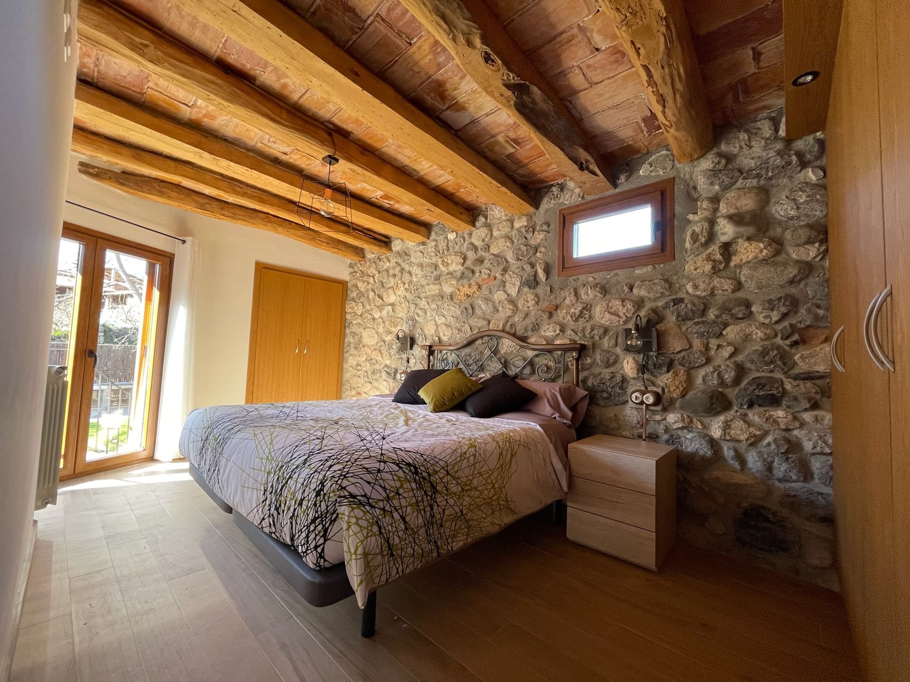 Habitació principal amb bigues de fusta, paret de pedra i sortida al jardí